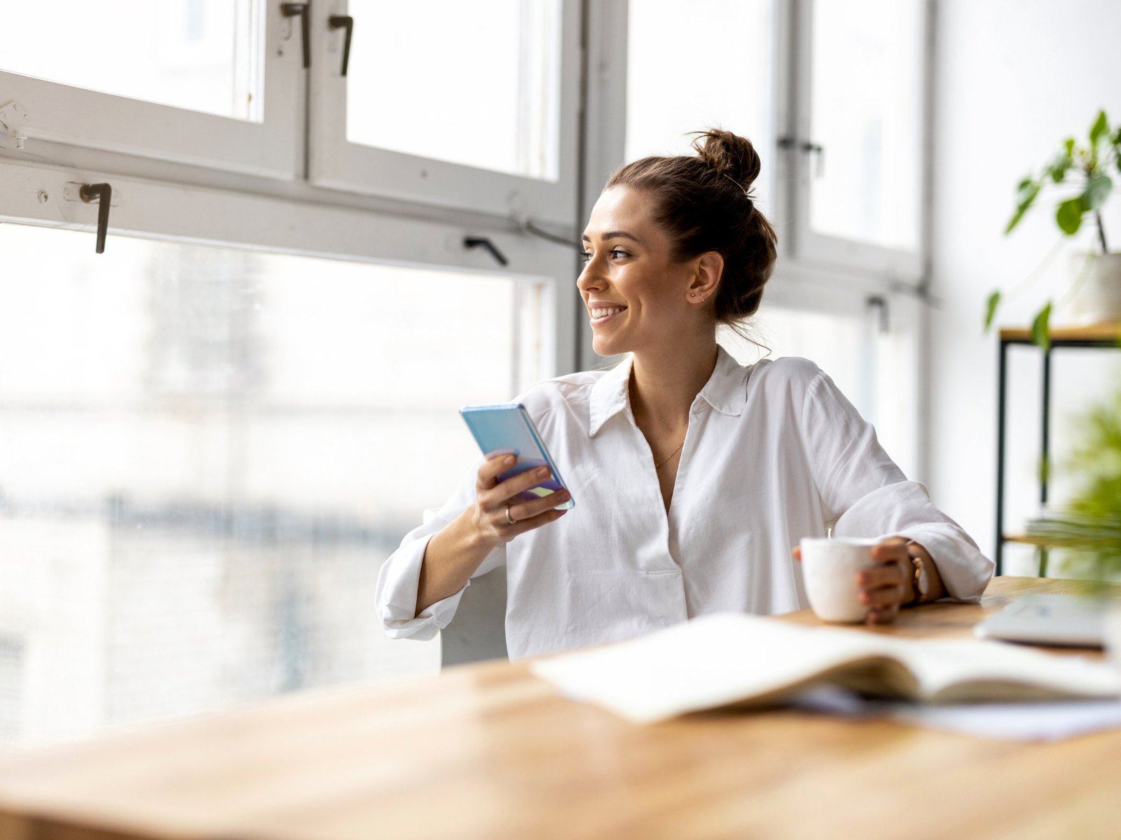 A business owner smiling at her desk by a bright window, phone in hand